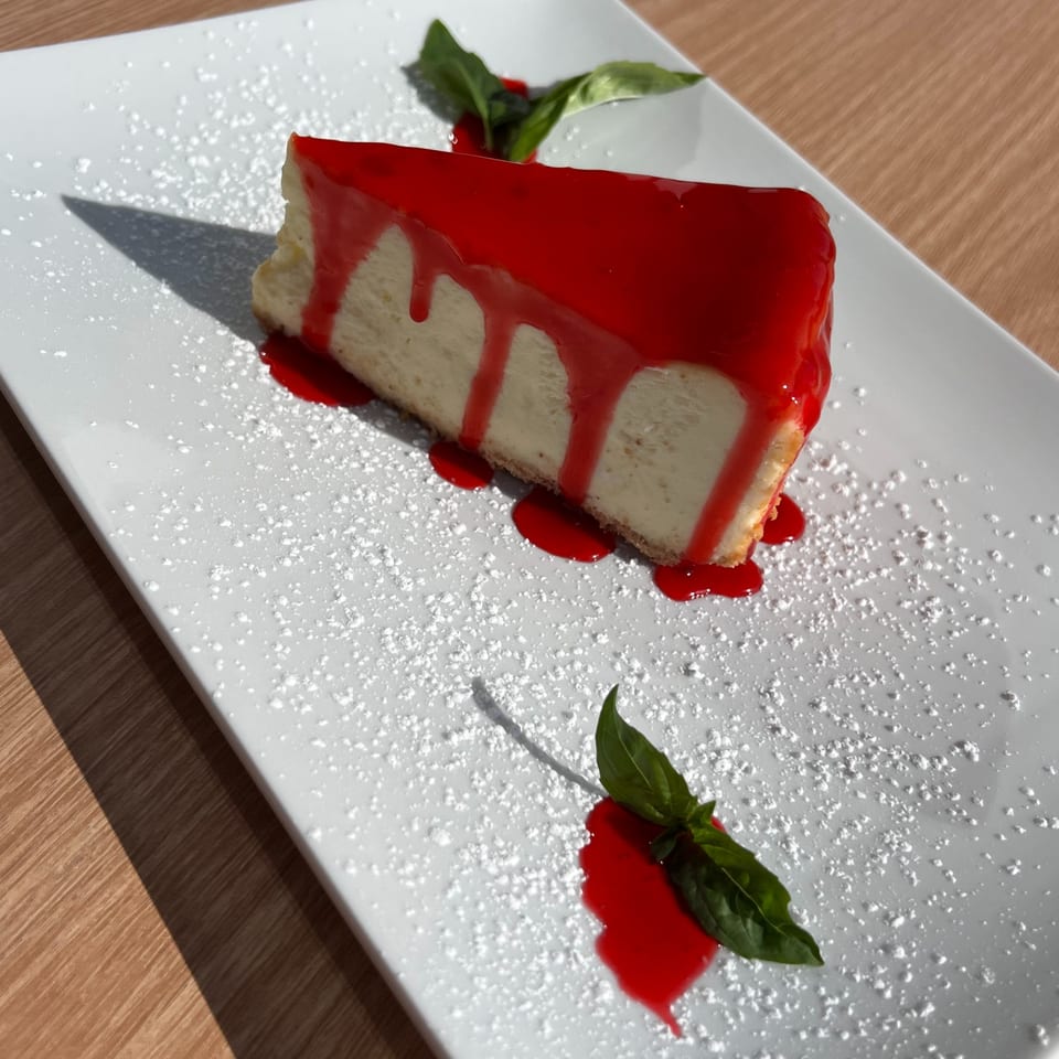 A slice of cheesecake with red raspberry sauce and powdered sugar on a white rectangular plate.