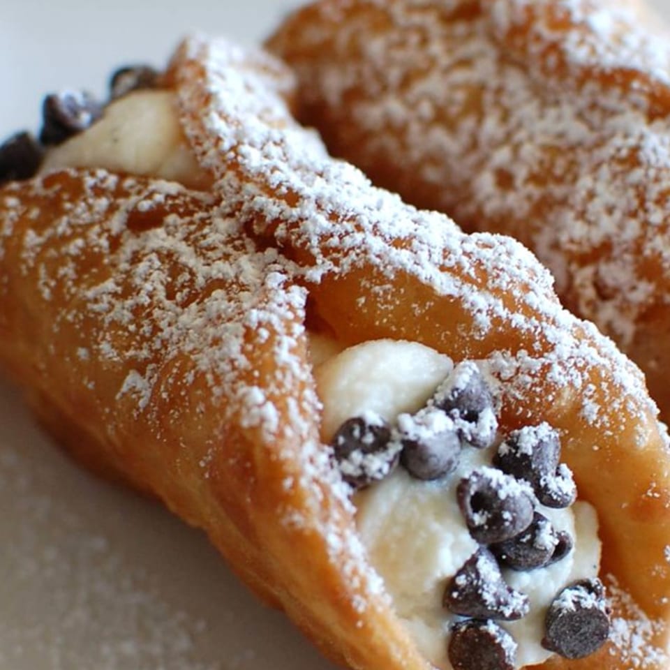 Two cannolis, one in focus, filled with cream and chocolate chips, dusted with powdered sugar.