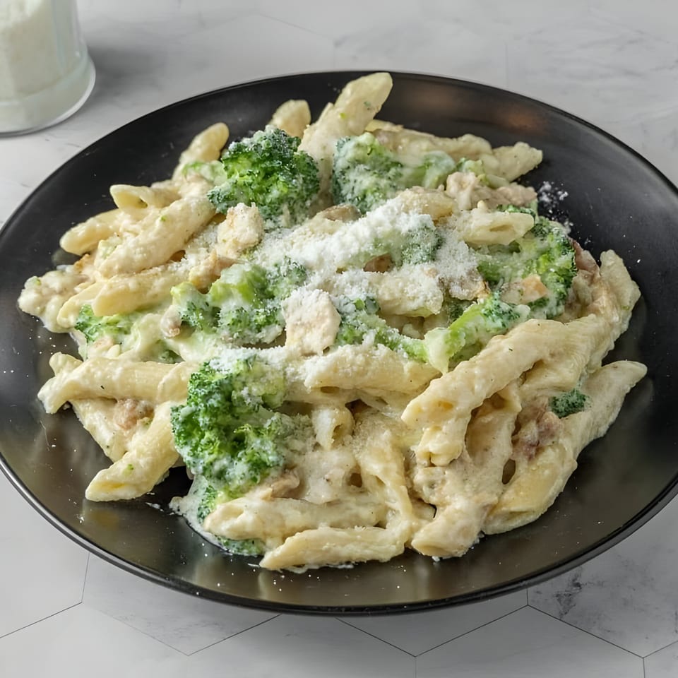 Creamy penne pasta with broccoli florets and grated Parmesan cheese on a black plate.