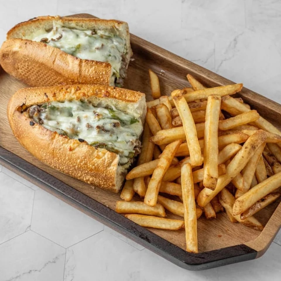 Cheesesteak sandwich with melted cheese and green peppers, served with a side of fries on a wooden tray.