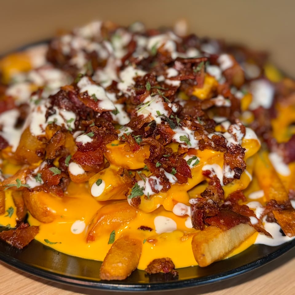Loaded fries with melted cheese, crispy bacon bits, and a drizzle of white sauce on a black plate.